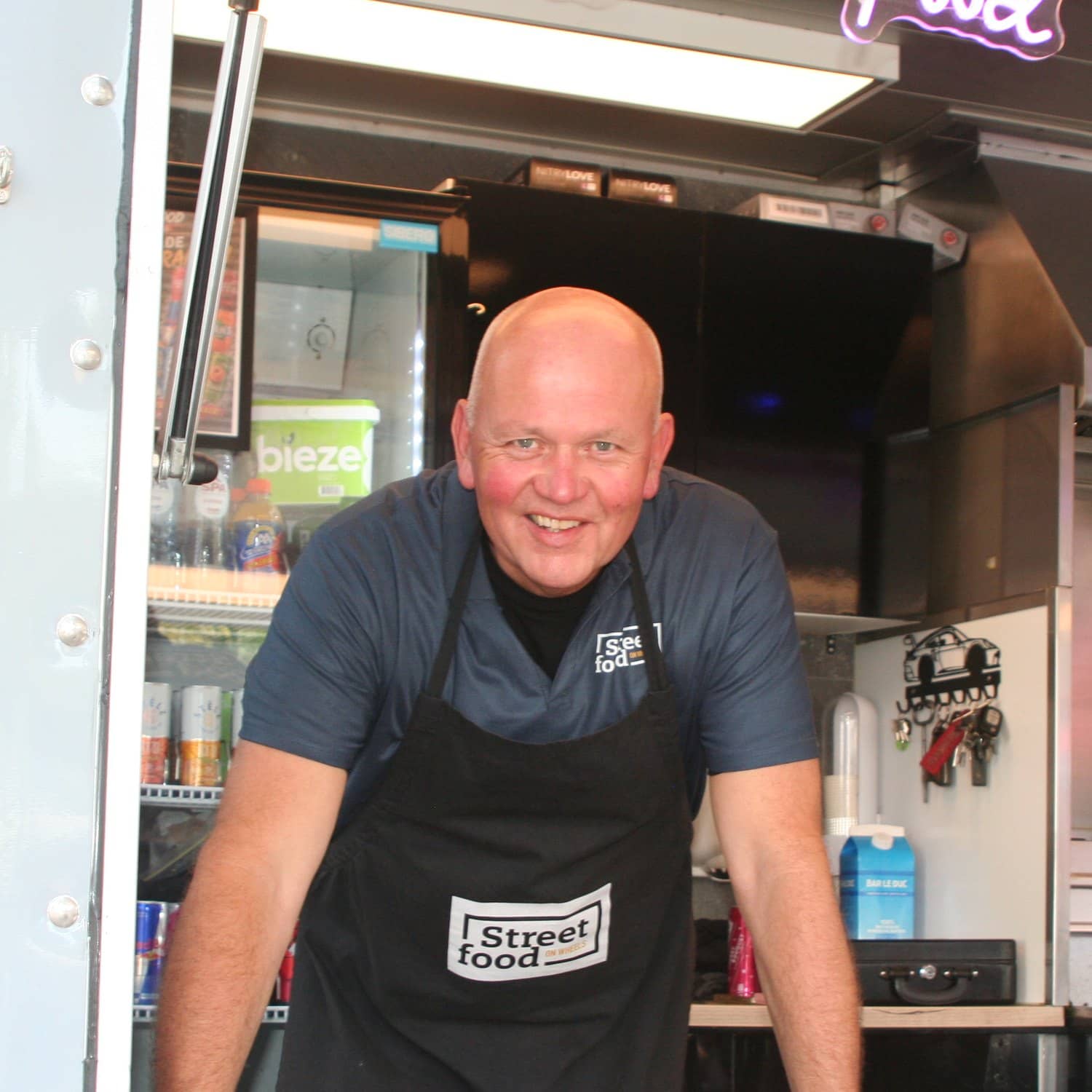 De eigenaar leunt lachend over de toonbank van de truck, in een schort met het logo