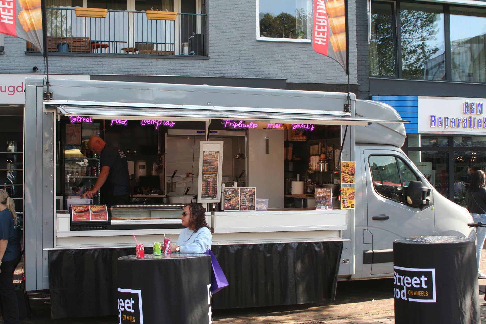 De zilveren foodtruck op de markt, met een klant aan de statafel met het logo